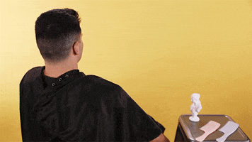 Video gif. Barber cape is cleanly snatched off from a man who just got a fresh, new haircut. He smirks confidently at himself in a small, pink handheld mirror, and two pairs of hands come from behind to stroke his head.