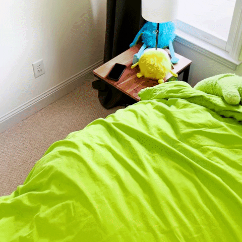 Video gif. Person in a giant smiley lime green mascot costume pulls back the lime green covers of their bed and stretches their arms out then side to side. 
