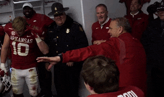 Sports gif. The Arkansas Razorbacks are in a team meeting in their locker room. The coach is revving up his fist and he jumps up and gives a big fist bump to rally the team. Everyone jumps with him and grin and whoop as they celebrate.