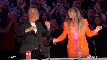 Reality TV gif. On America's Got Talent, Howie Mandel and Heidi Klum dance and clap their hands in time, standing up behind the judges' table, while the audience does the same.