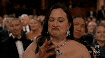 Oscars 2024 GIF. Lily Gladstone, seated at the Oscars, shrugs and nods, gesturing “so-so,” half-confirming.