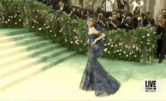 Met Gala 2024 gif. Zendaya poses in place, giving a full view of her her midnight blue and dark teal Maison Margiela tulle gown with a trumpet skirt covered in royal blue and emerald green diagonal stripes and fruit-like embellishments adorning one side of the waist and arm. 