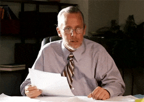 Video gif. Man waves his hands over paper documents on his desk expectantly, "What?"