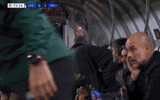 Video gif. Pep Guardiola sits on the sidelines of a soccer game. He has his hand by his chin with his pinky in his mouth as he looks around with a stressed out expression. He's remains still while players move all around him with a tense and lively energy.
