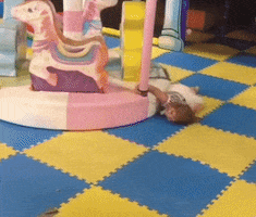Video gif. A little girl lies face down on the floor. With one hand, she clings to the pole of a small, rotating carousel, and is dragged in a circle.