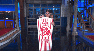 TV gif. Stephen Colbert has 3D glasses on and is holding a popcorn carton that's the size of his body. He hugs the carton and eats from the top of it, riveted by what he's watching.