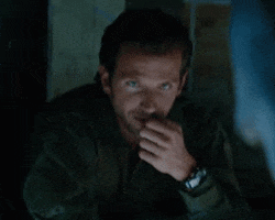 Celebrity gif. Bradley Cooper leans on a table and has his hand up to his lip. He looks at us flirtatiously and winks.