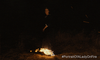 Movie gif. Adele Haenel as Heloise in Portrait of a Lady on Fire standing in a grassy clearing at night, with the bottom edge of her dress aflame.