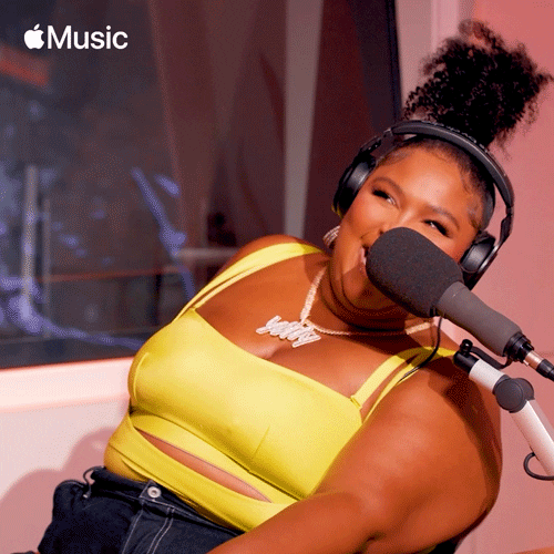 Celebrity gif. Lizzo leans forward and laughs as she wears headphones at a microphone. 