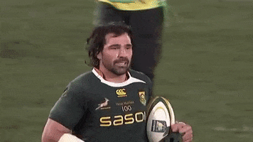 Sport gif. Victor Matfield, a South African rugby player, is on the field and he runs by holding the ball. He spots the cameras on him and he gives a closed lipped smile while waving his hand in the air.
