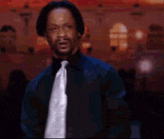 Celebrity gif. Wearing a black shirt with a white tie, Katt Williams looks around in confusion.