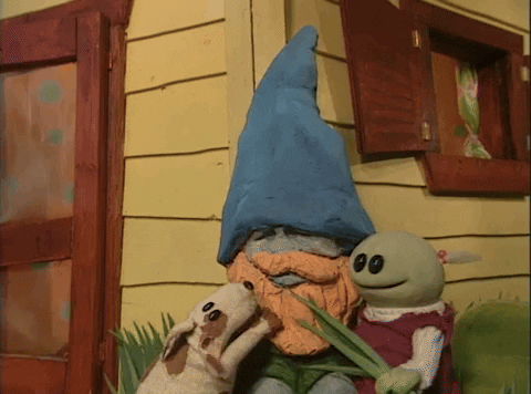 A green little girl and her brown dog are pestering a gnome that is sitting at the corner of what looks to be a yellow house.