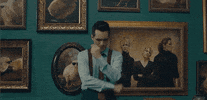 Music video gif. Brendon Urie is standing in a hallway filled with picture frames and he looks down, upset at  something. He sighs deeply and crosses his arms in contemplation.