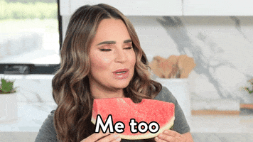 Video gif. Woman is holding a slice of watermelon that she's just taken a bite out of. She shakes her head and says, "Me too," while chewing.