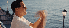 Movie gif. Leonardo DiCaprio as Jordan in Wolf of Wall Street. He's standing at the edge of a bay and he holds a stack of money which he tosses out, one bill at a time, and they fly in the wind.
