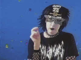 Video gif. A person in all black clothing and heavy goth makeup dons a "Happy New Year" mask and blows a party blower in front of a confetti background. Text, "New Year, same pain."
