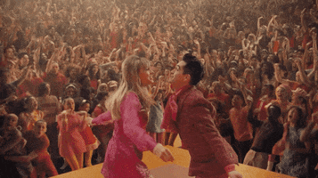 Music video gif. Taylor Swift and Brendon Urie in the Me music video stand up on stage shimming their shoulders towards each other. A crowd of people look up at them and dance. 