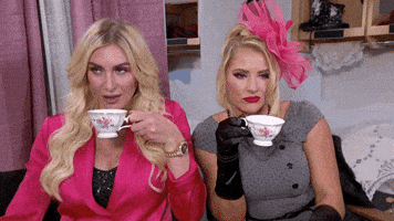Celebrity gif. Wrestlers Lacey Evans and Charlotte Flair wear fancy clothes while sipping tea from white cups. They look around with alluring eyes like they're ready for some juicy gossip.