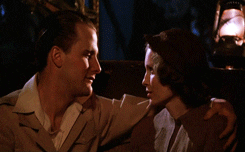 Movie gif. Jeff Daniels as Tom Baxter sits next to Mia Farrow as Cecilia in "The Purple Rose of Cairo," with his arm around her shoulder and leans in to kiss her.