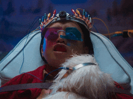 Movie or TV gif. Person lays back wearing purple and blue tinted protective glasses and a metal device on their head with colorful bulbs and wires coming out of it. They have something in their mouth as they try to speak. A person wearing a blue medical glove reaches in from out of frame to silence their speech.