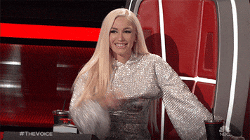 Reality TV gif. Gwen Stefani on The Voice sitting in the judge's chair, smiling, and pointing her finger at herself.