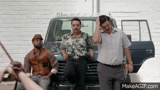 Video gif. Pablo Escobar puts a hand through his hair and walks toward us as two of his men lean against the hood of a car, watching.