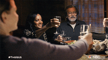 TV gif. Table full of characters on This is Us including Justin Hartley as Kevin and Griffin Dunne as Nick Pearson lean forward to clink their different glasses together to make a toast.