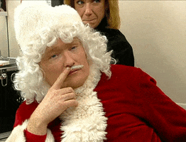 TV gif. Conan O'Brien is dressed as Santa Claus and he slowly spins in a chair while using one finger to stroke his thin mustache.