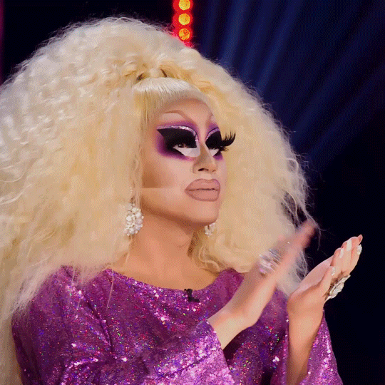 Reality TV gif. Trixie Mattel on Queen of the Universe, applauds as he nods to someone off screen. He wears full makeup, enormous eyelashes, blonde hair done up, and purple dress. 
