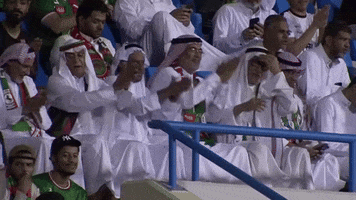 Sports gif. Saudi Arabian soccer fans cheer and clap in the stands. They all wear their traditional garb and sway in unison.