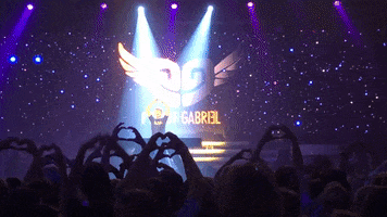 Video gif. Music artist Pieter Gabriel stands on stage in front of a cosmic backdrop with a twirling winged, heart-shaped logo. He interacts with an adoring crowd of fans, who are making hearts with their hands and recording him.