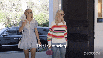 Reality TV gif. Paris Hilton in Paris in Love holds a small white dog wearing sunglasses and smiles as she enters through a large open door. Text, "This is amazing!"