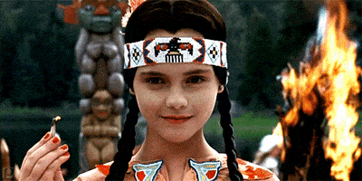 Movie gif. Christina Ricci as Wednesday Addams in The Addams Family. She's wearing a Native American headband and dress and she smiles sinisterly while holding a lit match. A fire rages behind her.
