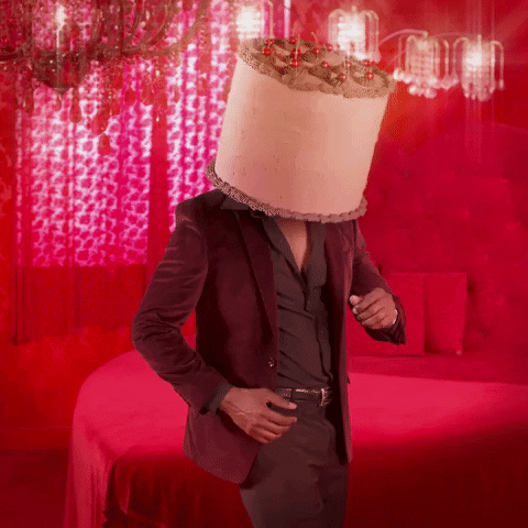 Video gif. A man in a burgundy suit is doing the two step while he wears a giant cake on his head.
