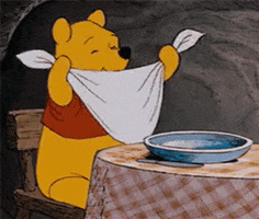 Cartoon gif. Winnie the Pooh sits in a wooden chair at a dinner table that has a large plate set in front of him. He ties a napkin around his neck, and then grabs the knife and spoon. He holds the utensils to signal he’s ready to eat and he wriggles in his seat with anticipation and excitement.
