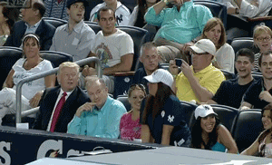 Video gif. Crowd at a baseball game does the wave. Several people pop up from their seats with their arms up and then sit back down. Donald Trump watches them and as the wave passes him, he raises his hand up a few seconds too late.