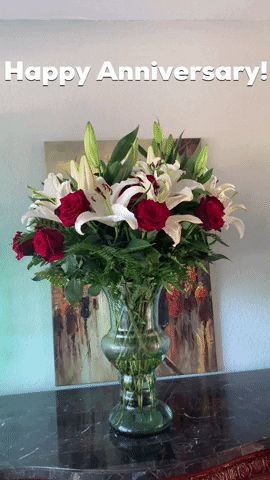 Video gif. A bouquet of white lilies and red roses in a glass vase with a painting obscured behind it. Text, "Happy Anniversary!"