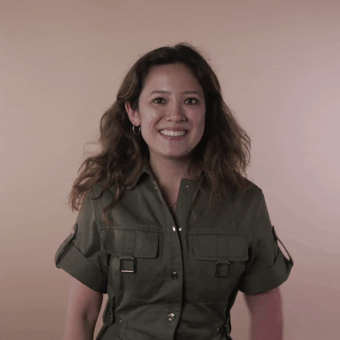 Reaction gif. A Disabled Vietnamese-American woman hemorrhagic stroke survivor with left-sided hemiplegia, brown sun-bleached hair styled in waves throws her arms around herself in a warm hug, smiling broadly.