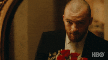 TV gif. Angus Cloud as Fezco in Euphoria wearing a suit and white collared shirt holds a bouquet of roses up to his nose as he stares at himself in the mirror, admiring his reflection.