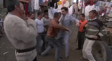Movie gif. Sacha Baron Cohen as Borat holds out his arms and shakes out some unique dance moves. He dances with a man in a group of other jolly men.