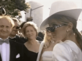 Celebrity gif. Celine Dion wears an abstract shaped hat that tilts over her eyes and a pair of sunglasses low on her nose. She poses, looking over her shoulder and over her sunglasses. People look at her smiling and nodding.