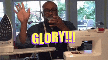 Video gif. Man is sipping a martini and has a sewing machine and iron board in front of him. He smiles and waves calmly as he sips. Text, "Glory!"