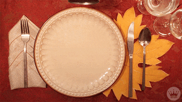 Stop motion gif. A plate surrounded by silverware with a playmat shaped like an orange leaf underneath.A hand holds a can of cranberry sauce and the sauce slides out and onto the plate. A piece of bread and corn transforms it into a pilgrim hat. The cranberry sauce pilgrim hat is replaced by a turkey made out of a oll, stuffing, corn, and carrots. The turkey flies away. 