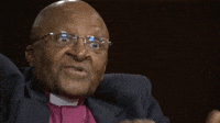 Video gif. Archbishop Desmond Tutu flashes excited eyes and seems to say Oh during an interview.