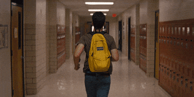 Movie gif. Miles Teller as Sutter Keely in The Spectacular Now runs down a dimly lit, narrow school hallway with a yellow backpack on.