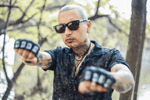Video gif. A heavily tattooed man wearing sunglasses holds out two gloved fists, punching his knuckles toward us.