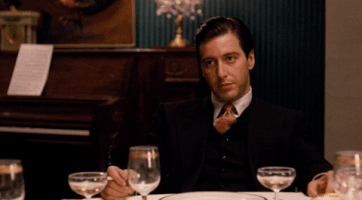 Movie gif. Al Pacino as Michael Corleone in The Godfather. He sits at a table and waits, as he stares at someone expectantly but absentmindedly fingers something on the table. A cigarette is lit next to him.