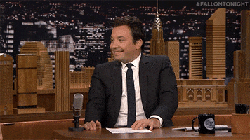 Tonight Show gif. Jimmy Fallon sits at his desk smiling and tapping his fingers expectantly as he raises his eyebrows.