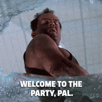 Movie gif. Bruce Willis as John McClane in Die Hard looking down through broken glass, saying, "welcome to the party, pal."
