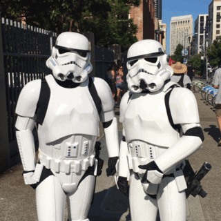 Star Wars gif. Two people dressed as Storm Troopers give a simultaneous thumbs up at us, standing on a city sidewalk.